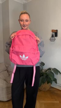 Mochila rosa da Adidas com um logótipo branco, bolso frontal com fecho e alças ajustáveis. O material parece durável e suave.