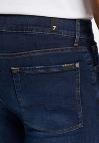 Jeans in denim blu scuro con cuciture arancioni, una tasca posteriore con etichetta nera e una superficie liscia e textured.