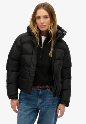 Vrouw in een zwarte pufferjack met capuchon, zwarte gebreide trui, gevlochten bruine riem en blauwe jeans, staande voor een witte achtergrond.