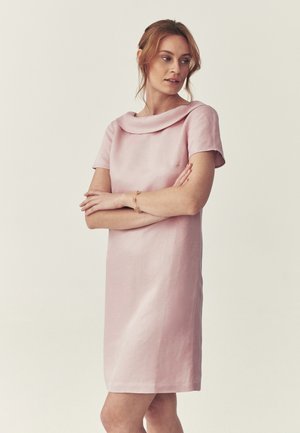 Donna con capelli castano chiaro, vestita con un abito rosa a maniche corte con un ampio colletto, braccia incrociate, che guarda di lato, su uno sfondo chiaro e uniforme.