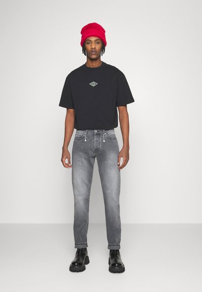T-shirt preta com logótipo verde, jeans cinzentos ajustados com detalhes em zíper e botas pretas de plataforma. Gorro de malha vermelho na cabeça.