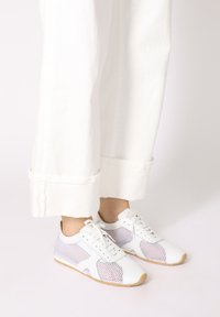 Weiße Sneaker mit Netzpaneelen, Schnürdesign, strukturiertem Gummisohlen und kontrastierenden hellgrauen Akzenten. Getragen mit hochgerollten weißen Hosen.