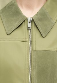 Κοντινό πλάνο σε ελιά-πράσινο μπουφάν με σουέντ γιακά και μπαλώματα, με ασημένιο μεταλλικό φερμουάρ μερικώς κλειστό στο κέντρο μπροστά.