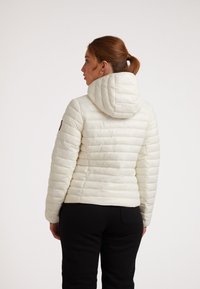Giacca a vento bianca con texture trapuntata orizzontale, cappuccio aderente e bordo a costine. Indossata con pantaloni neri, vista da dietro.