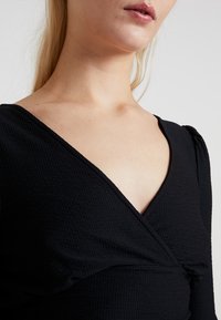 Schwarzes, strukturiertes Oberteil mit tiefem V-Ausschnitt, gerafftem Detail in der Taille und langen Ärmeln mit dezenten puffierten Akzenten.