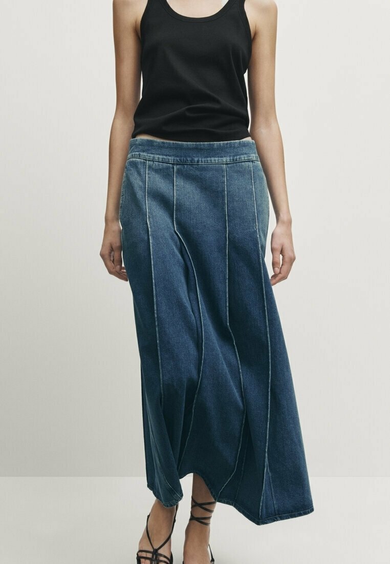 Personne portant un haut noir sans manches, une longue jupe en jean bleu évasée avec des coutures verticales et des chaussures noires à brides, debout devant un fond uni.