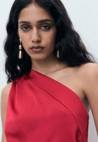 Red, one-shoulder dress in smooth fabric. Features a draped design and a defined neckline. Gold earrings with cylindrical accents.