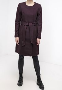 Kabát z burgundské vlněné směsi s dlouhými rukávy, kulatým výstřihem, zúženým pasem a bočními kapsami. Nosený přes černé texturované kalhoty a boty.