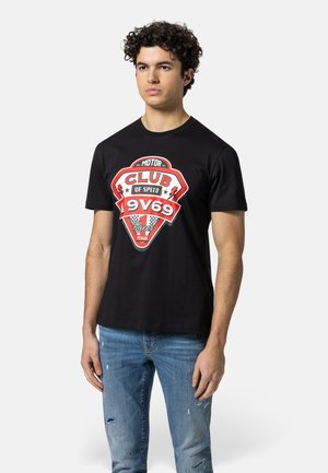 Junger Mann mit lockigem Haar, der ein schwarzes T-Shirt mit einem roten und weißen "Club of Speed 9V69 Racer Italia"-Logo und zerrissene blaue Jeans trägt.