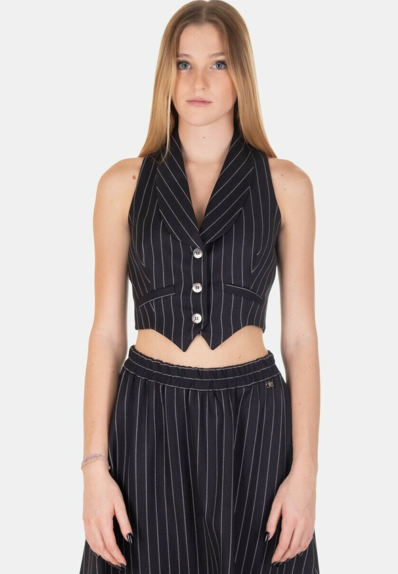 Gilet corto a righe sottili bianche e nere con colletto a revers, cinque bottoni e due tasche frontali. Indossato con una gonna abbinata.
