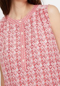 Blusa senza maniche a righe rosse e bianche con scollo rotondo, caratterizzata da motivi floreali e una chiusura frontale con bottoni.