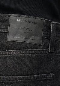 Μαύρο τζιν από denim με μαύρο δερμάτινο ταμπελάκι με την ένδειξη "JACK & JONES" πάνω από την πίσω τσέπη, φορεμένο με μαύρο ριμπ μπλουζάκι.