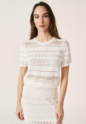 Mujer de cabello largo castaño, vestida con un top blanco de manga corta tejido y una falda a juego, de pie frente a un fondo beige liso.