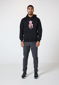Schwarzer Hoodie mit einem grafischen Druck eines Bärencharakters in Pink und Braun, kombiniert mit grauen Cargo-Hosen und schwarzen Turnschuhen.