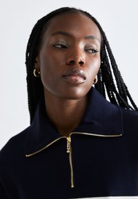 Sweatshirt blu navy con zip e colletto alto, caratterizzato da una cerniera dorata e dettagli a strisce bianche. Texture liscia, design aderente.