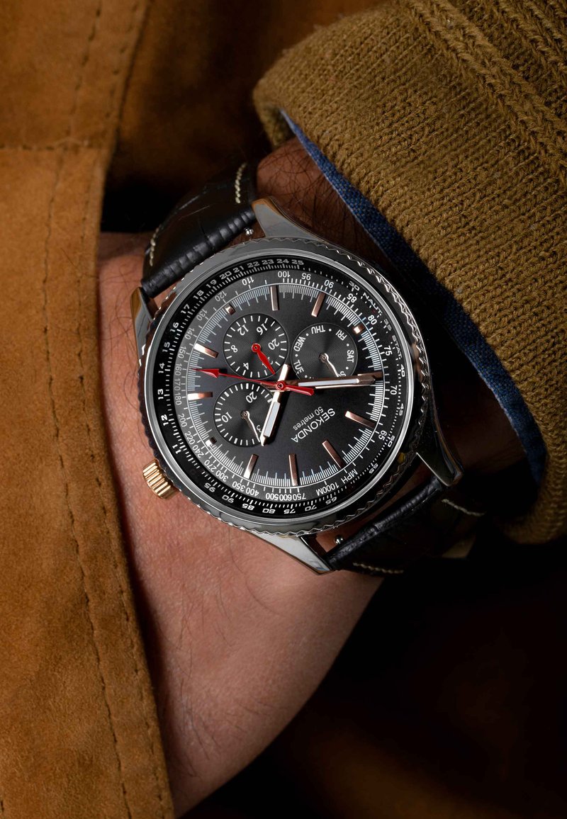Black chronograph watch with silver accents, textured bezel, and leather strap. Features three sub-dials and red hands on a black face.