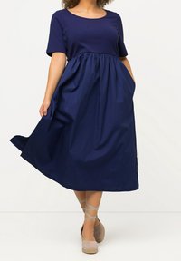 Mörkblå klänning med korta ärmar, en figurnära bodice och en utsvängd kjol. Har en slät yta och sidofickor.