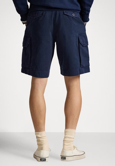 Tumši zilas kravas īsās bikses ar sānu kabatām, izgatavotas no izturīga auduma, ar gludu tekstūru un ceļgala garuma dizainu; kombinētas ar baltām sporta kurpēm.