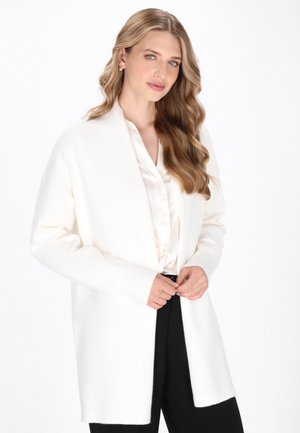Witte cardigan met open voorkant, geribbelde manchetten en een ontspannen pasvorm. Gedragen over een lichte satijnen blouse en zwarte broek. Zachte textuur zichtbaar.