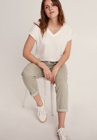 Weißes V-Ausschnitt-T-Shirt, kurze Ärmel, kombiniert mit hochgekrempelten, hellgrünen Hosen. Getragen mit weißen Turnschuhen, sitzend auf einem weißen Stuhl.