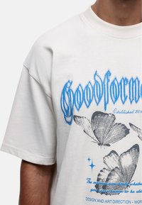 Witte katoenen t-shirt met blauwe grafische tekst "Goodform" en grijze vlinderillustraties, met een ronde hals en korte mouwen.