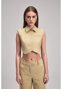Korte mouwloze blouse van beige stof met een spitse kraag, voorzien van een knoopsluiting en een klein horizontaal pocketdetail.
