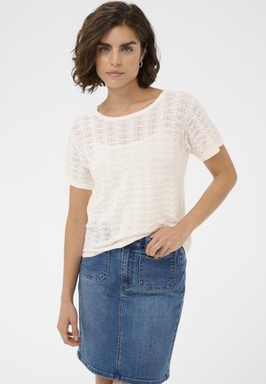 Naine kannab valget lühikeste varrukatega kootud pluusi ja põlveni ulatuvat sinist teksaseelikku, seistes ühe käega taskus.