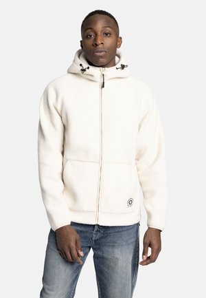 Sudadera blanca de forro polar con cremallera, capucha ajustable y dos bolsillos frontales. Presenta un logo circular en la parte inferior derecha. Combinada con jeans azules.