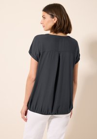 Blusa corta de manga corta gris oscuro con un dobladillo redondeado, fruncida en la cintura, que presenta un sutil pliegue en la parte trasera y una textura de tela suave.