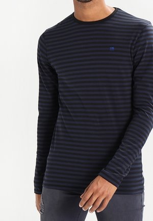 Hombre con camisa de manga larga a rayas horizontales en azul marino y negro y pantalones oscuros, de pie con una mano cerca de su muslo.