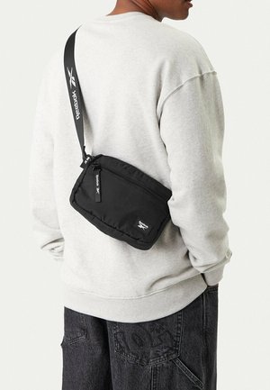 Person wearing light gray sweatshirt and dark jeans with black Reebok crossbody bag slung over their back.