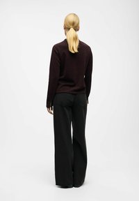 Persoon met een blonde paardenstaart, gekleed in een donker lange mouw top en wijde zwarte broek, staand tegenover een effen witte achtergrond.