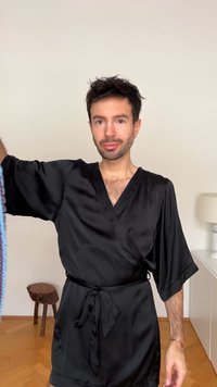 Melns sātna halāts ar apkakli, platām piedurknēm un jostu viduklī. Fonā redzama gaismas krāsas mēbeles un koka grīda.