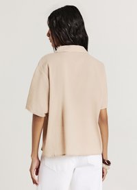 Chemise beige ample en tissu texturé, avec col et manches courtes, associée à un pantalon blanc et un accessoire cheveux simple.