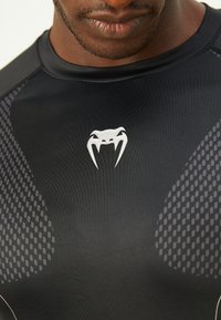 Gros plan sur une personne portant un t-shirt de sport noir avec des panneaux à motifs gris et un logo stylisé de visage avec des dents blanches sur la poitrine.