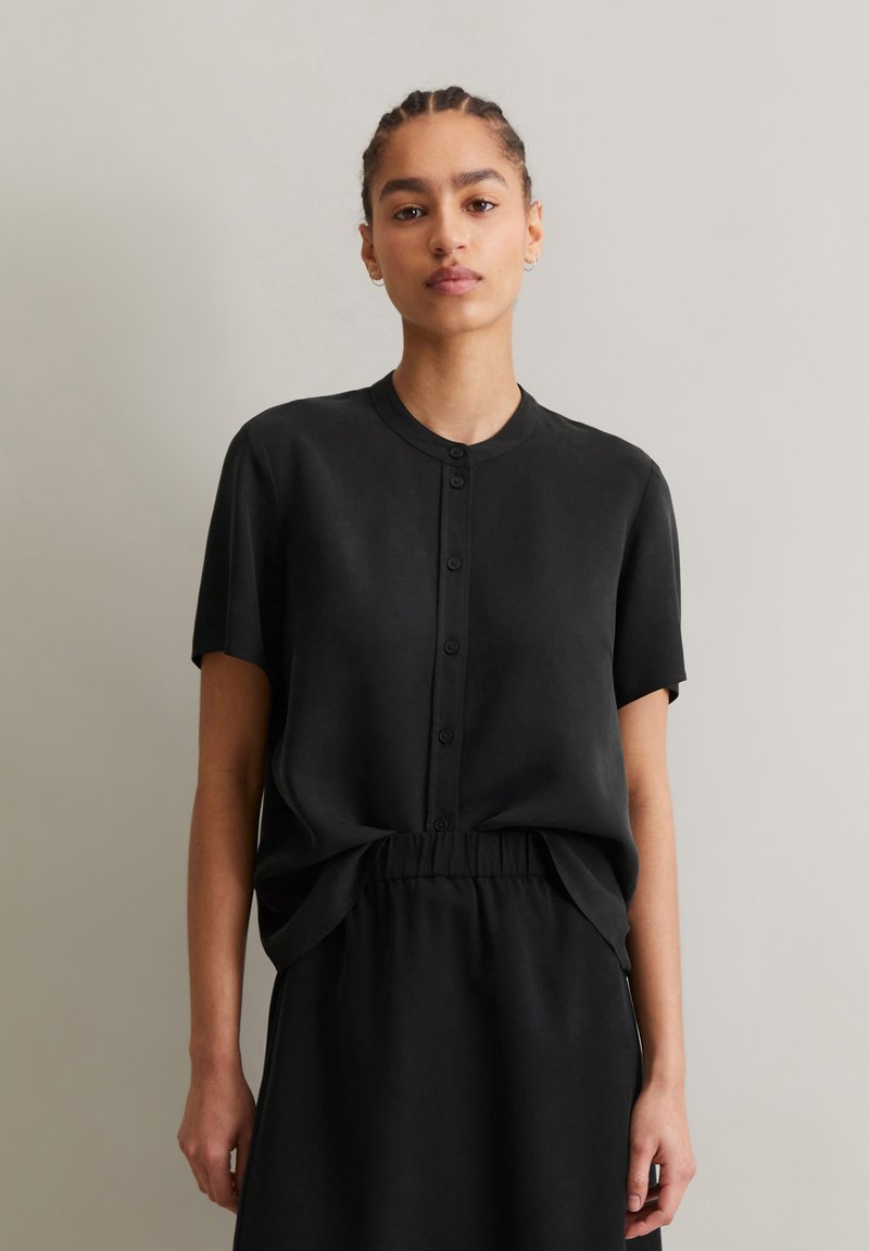 Schwarzes Kurzarm-Hemd mit Knopfleiste, aus glattem Stoff. Verfügt über einen Rundhalsausschnitt und einen lockeren Schnitt, kombiniert mit einem passenden Rock.