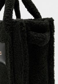 Sac en tissu noir texturé avec une surface douce, doté de deux poignées et d'un panneau en cuir noir contrastant avec des détails en matériel.