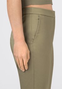 Boční pohled na osobu oblečenou v olivově zelených kalhotách s vysokým pasem a zkráceném olivově zeleném topu, s pravou rukou uvolněně položenou na boku.