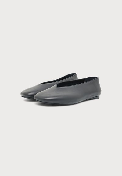 Paire de chaussures plates en cuir noir à enfiler avec un devant bas et des bouts arrondis sur un fond clair uni.