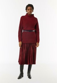 Burgundowy dzianinowy sweter z kołnierzem typu turtleneck w zestawie z dopasowaną plisowaną spódnicą, przewiązaną w talii, uzupełnioną czarnymi botkami na obcasie.