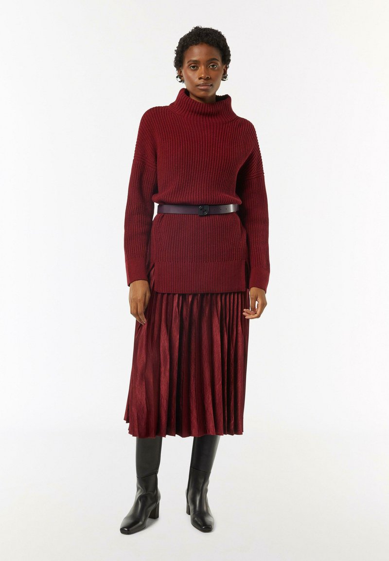Burgundowy dzianinowy sweter z kołnierzem typu turtleneck w zestawie z dopasowaną plisowaną spódnicą, przewiązaną w talii, uzupełnioną czarnymi botkami na obcasie.
