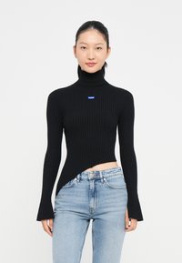 Maglione nero a collo alto a coste con un orlo asimmetrico. Presenta maniche lunghe e un logo blu. Abbinato a jeans sfrangiati blu chiaro.