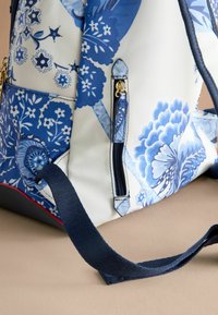 Mochila de patrón floral azul con un bolsillo con cremallera, hecha de tela duradera. Presenta una correa sólida de color azul oscuro y un acento rojo en la base.