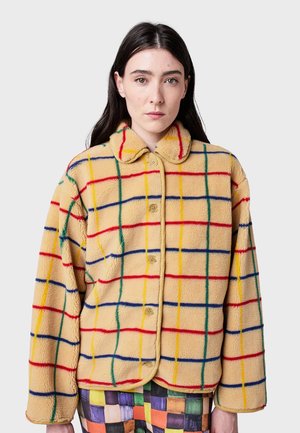 Vrouw met lang, donker haar, gekleed in een beige fleece jack met knopen en een veelkleurig ruitpatroon, en kleurrijke patchworkbroek.