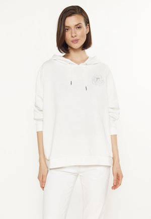 Vrouw met stijl bruin haar die een witte hoodie draagt met een klein bloemmotief op de borst en een witte broek tegen een effen achtergrond.
