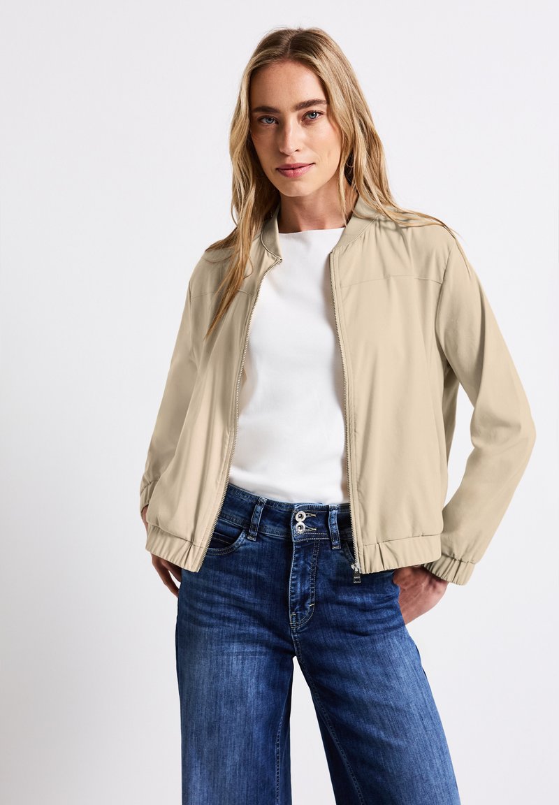 Chaqueta bomber beige con cremallera frontal, puños y dobladillo acanalados, usada sobre una blusa blanca y combinada con unos pantalones de pierna ancha azul oscuro.