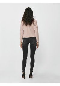 Femme aux cheveux bouclés portant un blazer rose clair, un jean skinny noir et des talons hauts noirs, debout de dos sur un fond blanc.
