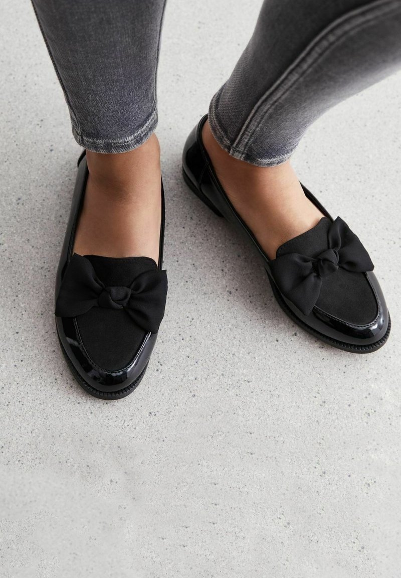 Pieds en mocassins noirs avec de grands nœuds en tissu, portés avec un jean skinny gris sur un sol gris clair tacheté.