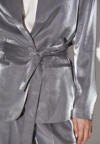 Zilveren satijnen blazer met een striktaille, lange mouwen en zakken. Heeft een gladde textuur en een lichte glans.