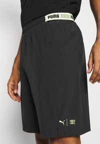 Mustat urheilushortsit, joissa on neonkeltaiset "PUMA"-vyötärönauha sekä "Puma"- ja "First Mile" -logot helman lähellä, käytössä olevan henkilön päällä.